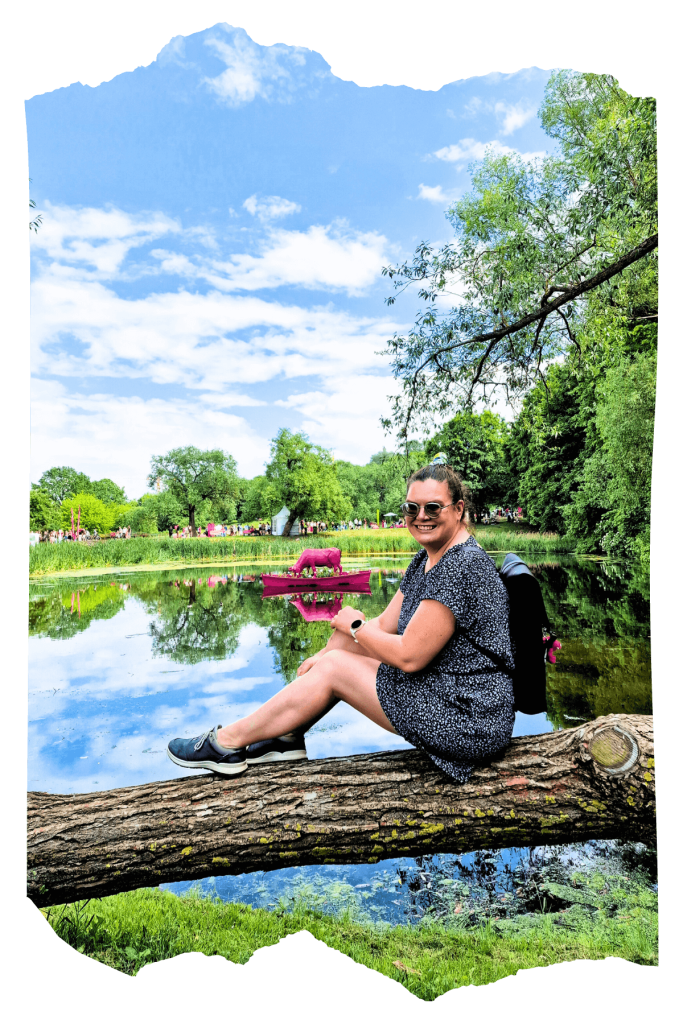 Ieva sitting on a fallen tree by a pond, her legs bent and resting on the trunk with her hands on her knees. In the middle of the pond there is a pink cow in a pink boat as a decoration, and the clear sky with a few clouds is reflected in the water.