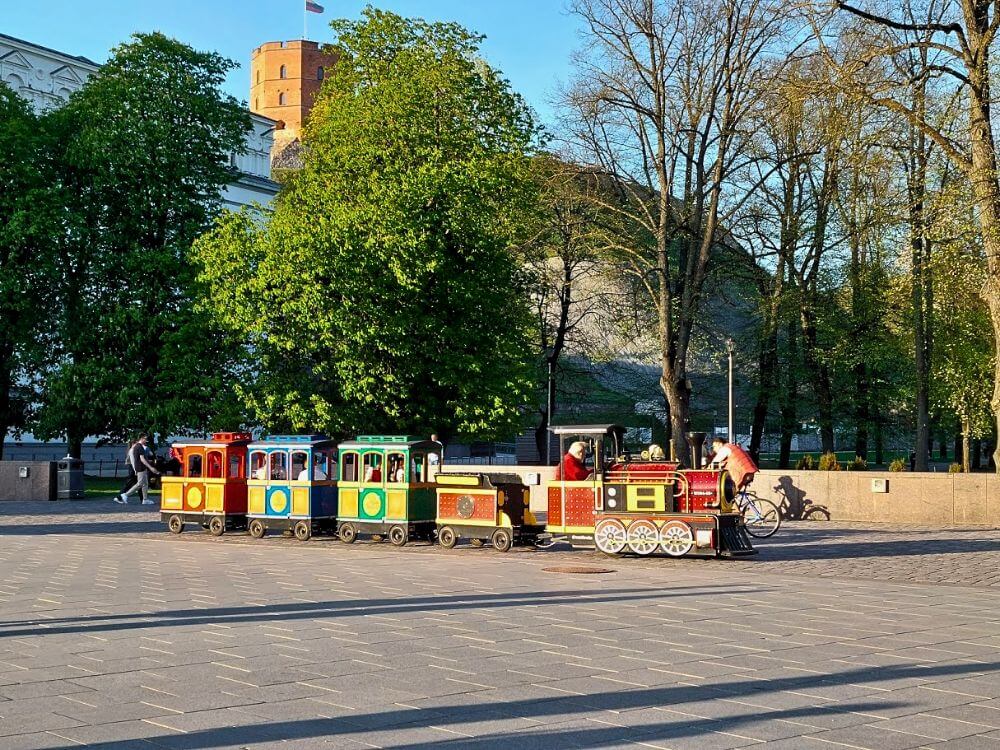 A colorful children’s train crossing Cathedral Square in Vilnius with Gediminas Castle Tower visible behind the trees.
