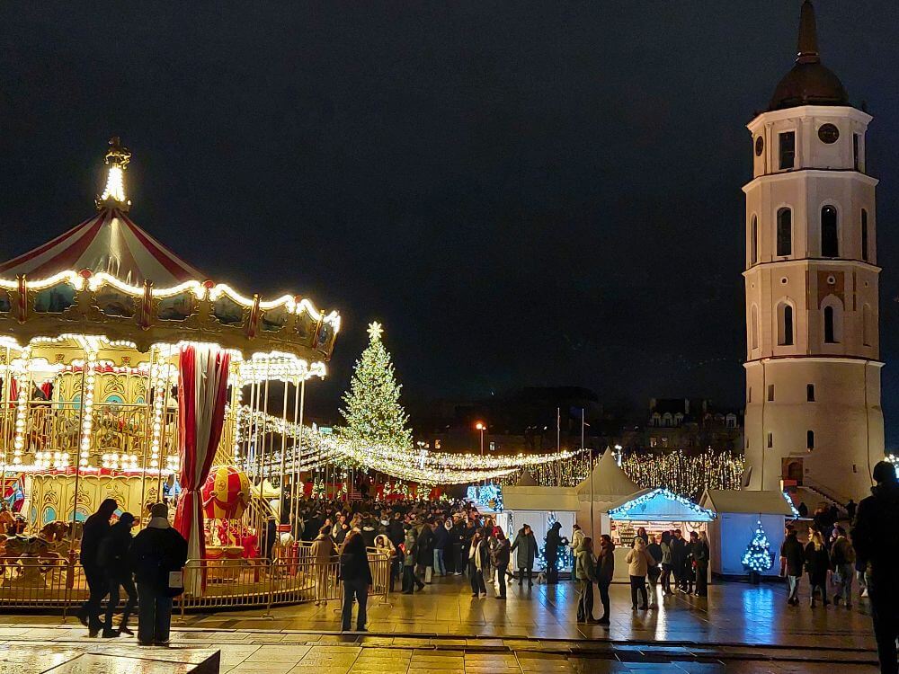Cathedral Square in Vilnius in December with a lit carousel, Christmas tree, market stalls, crowds, and the bell tower at night.