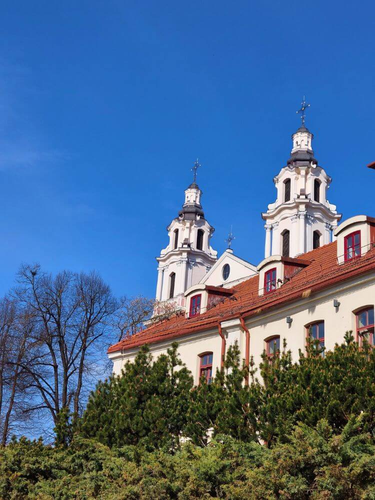 Church of St. Archangel Raphael in Vilnius rising above red rooftops and green shrubs under a clear blue April sky.