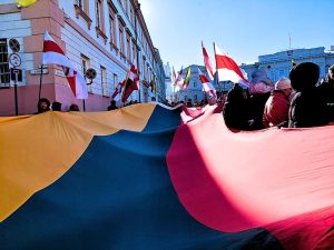 A large Lithuanian flag is carried through a crowd in the Presidential Palace courtyard during a February 16 celebration in Vilnius. People hold Lithuanian and red and white flags in bright winter sunlight.