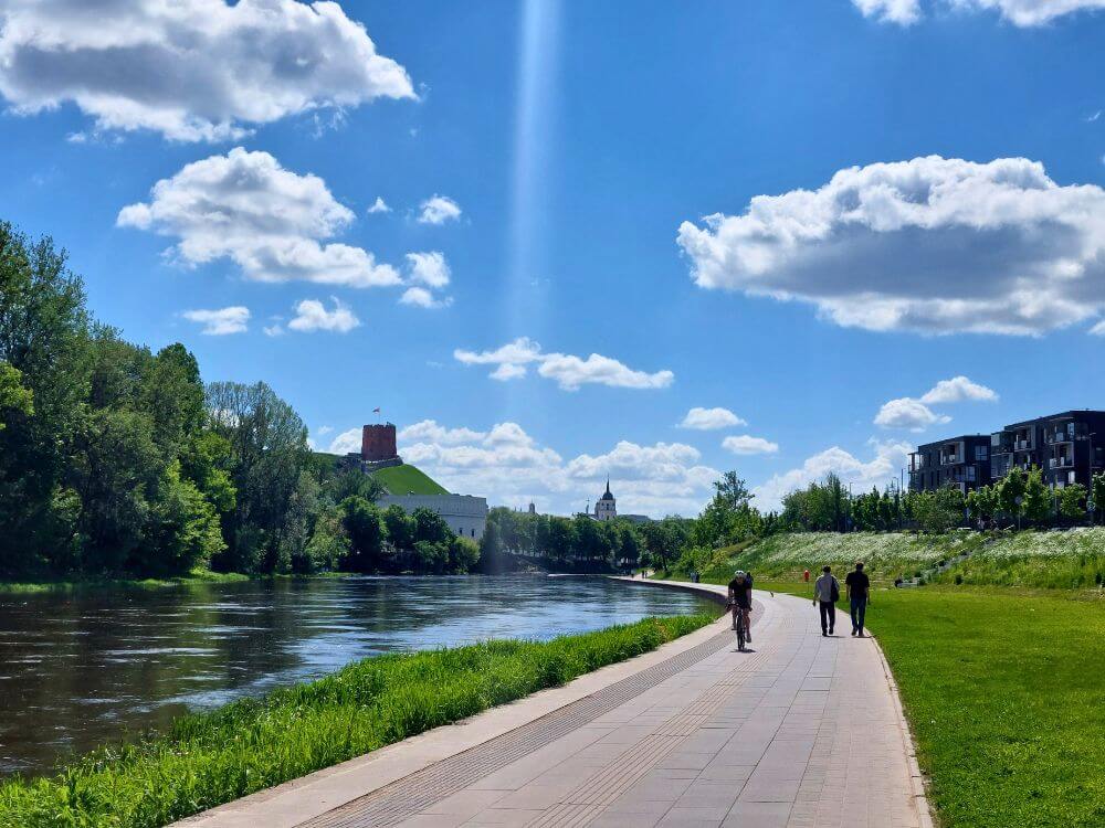 Neris riverfront in Vilnius in May with a riverside path, cyclists, walkers, Gediminas Castle Tower, and a sunny blue sky.