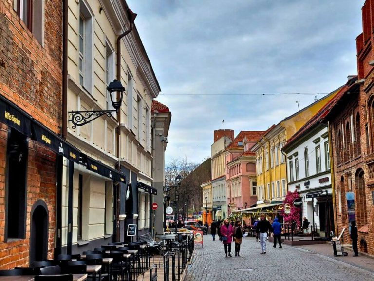Pilies Street in Vilnius Old Town with cobblestone pavement, outdoor cafe seating, colorful historic buildings, and Gediminas Castle Tower visible above the rooftops in the distance.