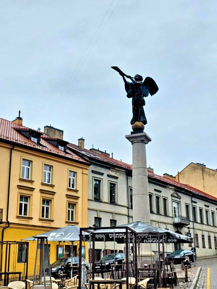 The Užupis Angel statue stands on a tall column above a cobblestone square in Vilnius. Pastel buildings, cafe umbrellas, and a gray sky surround the monument.