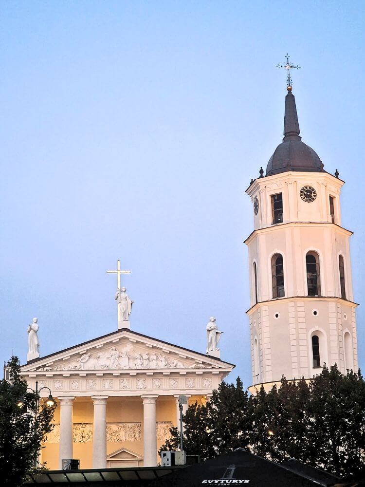 Vilnius Cathedral and its white bell tower glow at dusk under a pale blue sky. The cathedral roof has statues and a gold cross while dark trees frame the lower edge.