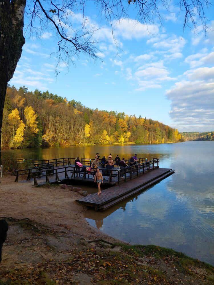 Žalieji ežerai (Green Lakes) in October with people on a wooden dock, calm water, and autumn trees along the shoreline.