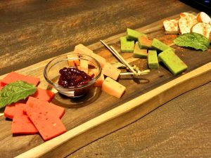 A wooden cheese board at Craft&Draft in Vilnius with red, yellow, green, and white cheese, berry jam, mint leaves, and small skewers.