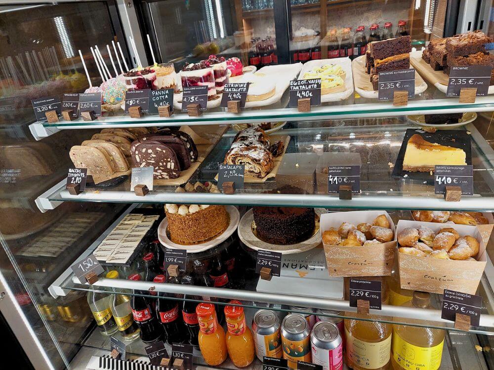Crustum bakery display case in Vilnius filled with cakes and tarts and brownies and pastries and doughnuts and bottled drinks. Visible labels include “Panna Cotta” and “Citrinine tartalete” and “Egzot. vaisių tartalete” and “Brownie su maskarpone” and “Brownie” and “Šimtalapis” and “Napoleono torto gabalėlis” and “Baskų sūrio pyrago gabalėlis”.