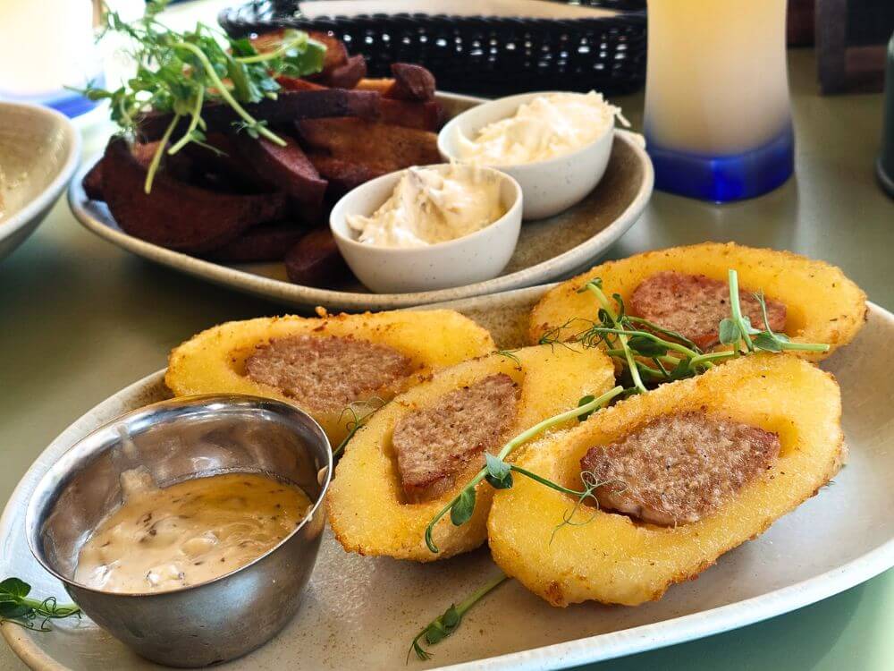 Fried cepelinai filled with meat at Katpėdėlė in Vilnius served with creamy sauce and pea shoots. A side plate of fried dark bread with two spreads sits in the background.