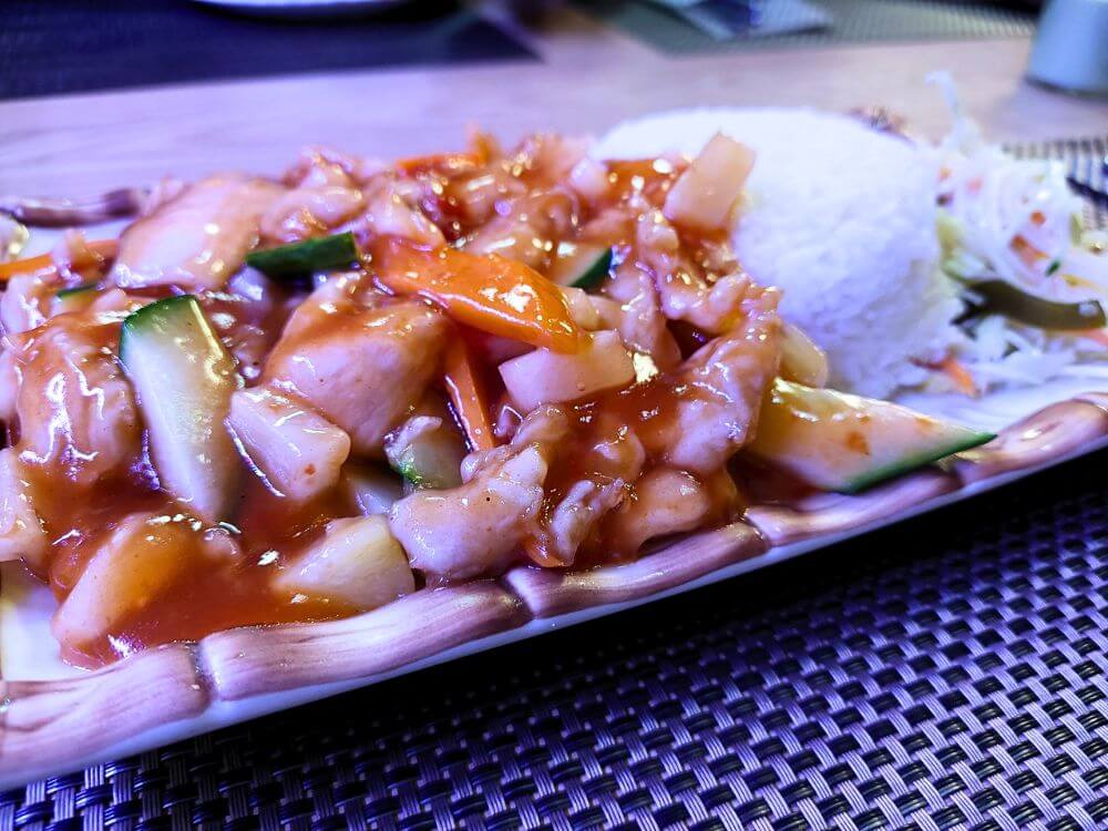 Chinese style chicken in glossy sweet and sour sauce at Kinų broliai in Vilnius, served with white rice and a small shredded cabbage salad on a rectangular plate.
