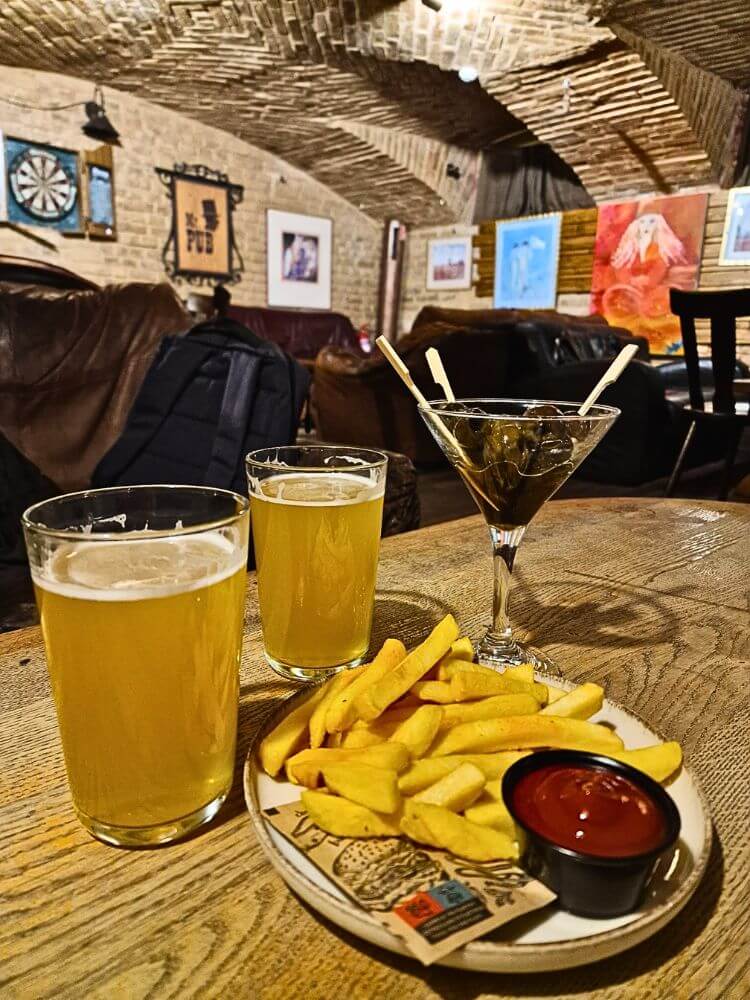 Two glasses of wheat beer sit on a wooden table at Mr. Pub in Vilnius beside a martini glass with olives and fries with ketchup. The cozy pub interior has brick arches with leather couches and framed art and a dartboard in the background.