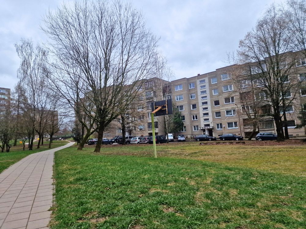 A sidewalk running through Justiniškės with apartment blocks, parked cars, bare trees, grass, and a small basketball hoop.