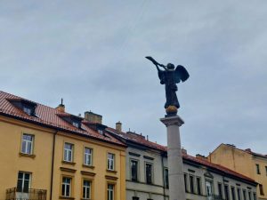 The Užupis Angel statue standing on a tall column above colorful old buildings and red tiled roofs in Vilnius.