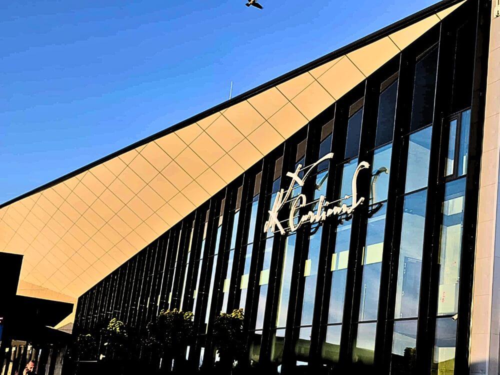 The modern glass exterior of Vilnius Airport with a “Departures” sign on the building and a clear blue sky above.