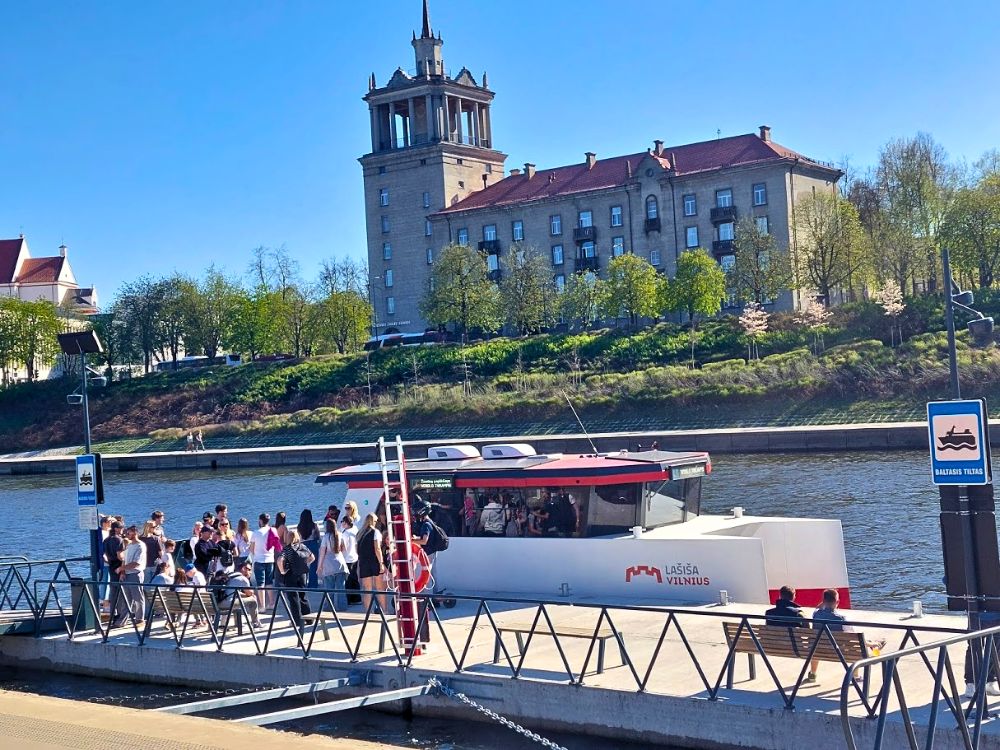 A white and red Vilnius public transport boat waits at a dock while passengers board beside the river. Historic buildings and green trees rise on the opposite bank under a clear blue sky.