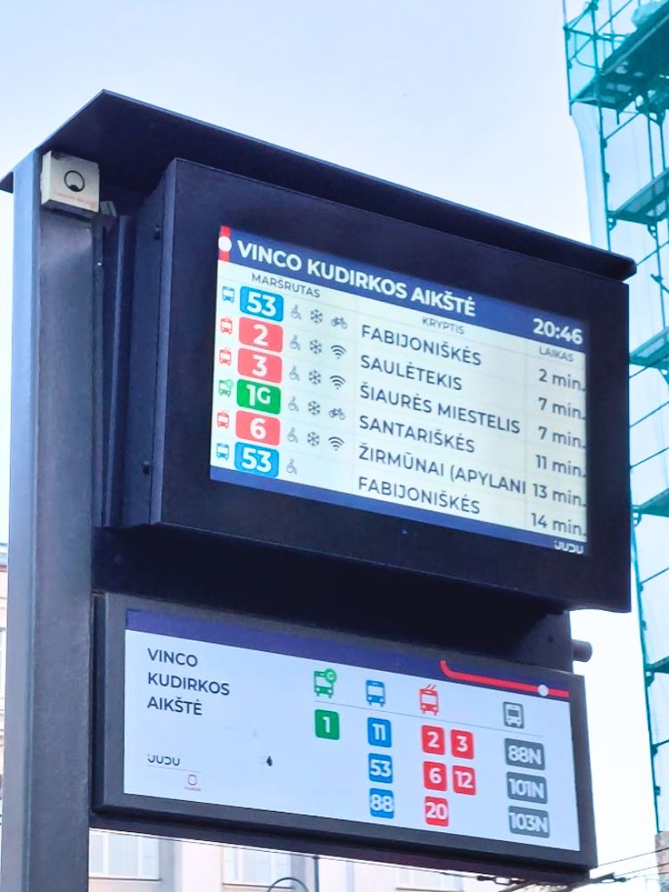 A digital arrival screen at the "Vinco Kudirkos aikštė" stop shows upcoming Vilnius public transport routes and wait times. The screen lists 53 to "Fabijoniškės" in 2 minutes. 2 to "Saulėtekis" in 7 minutes. 3 to "Šiaurės miestelis" in 7 minutes. 1G to "Santariškės" in 11 minutes. 6 to "Žirmūnai" in 13 minutes. and 53 to "Fabijoniškės" in 14 minutes.