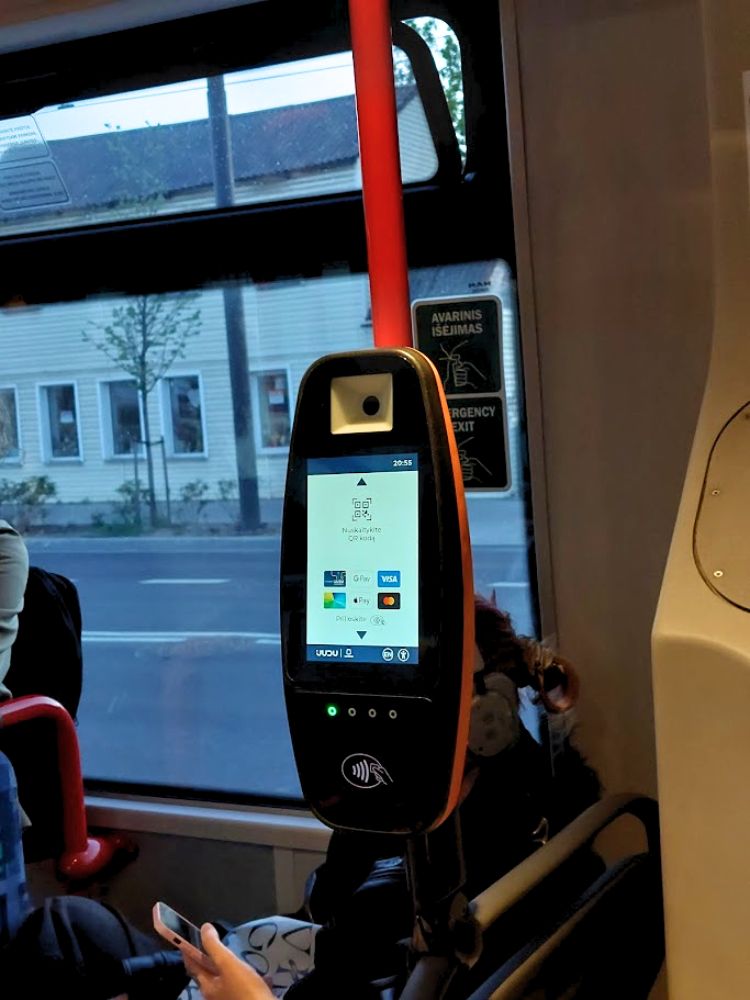 A ticket validator inside a Vilnius public transport bus beside a red handrail and window. The screen shows contactless payment options for buying or validating a ticket with a card or smart device.