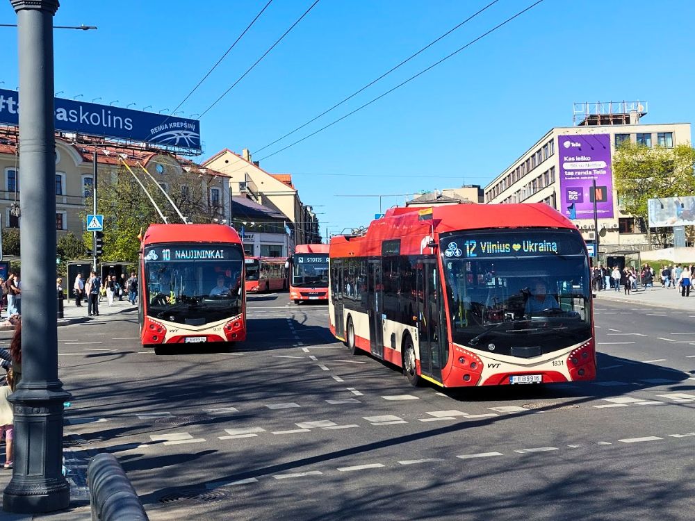 Two red trolleybuses marked 10 and 12 and a bus marked 53 move through a busy Vilnius street. Another bus turns into the stop in the background as pedestrians wait nearby.