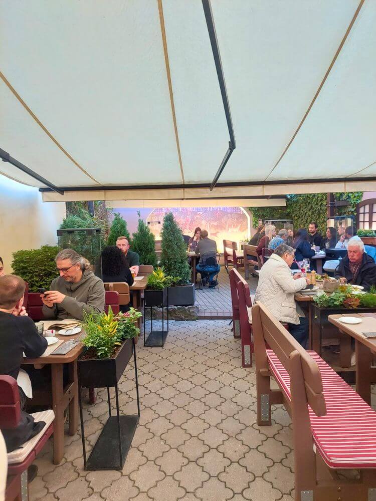 People dining on the covered terrace at Lokys restaurant in Vilnius with wooden tables, benches, planters, and courtyard walls.