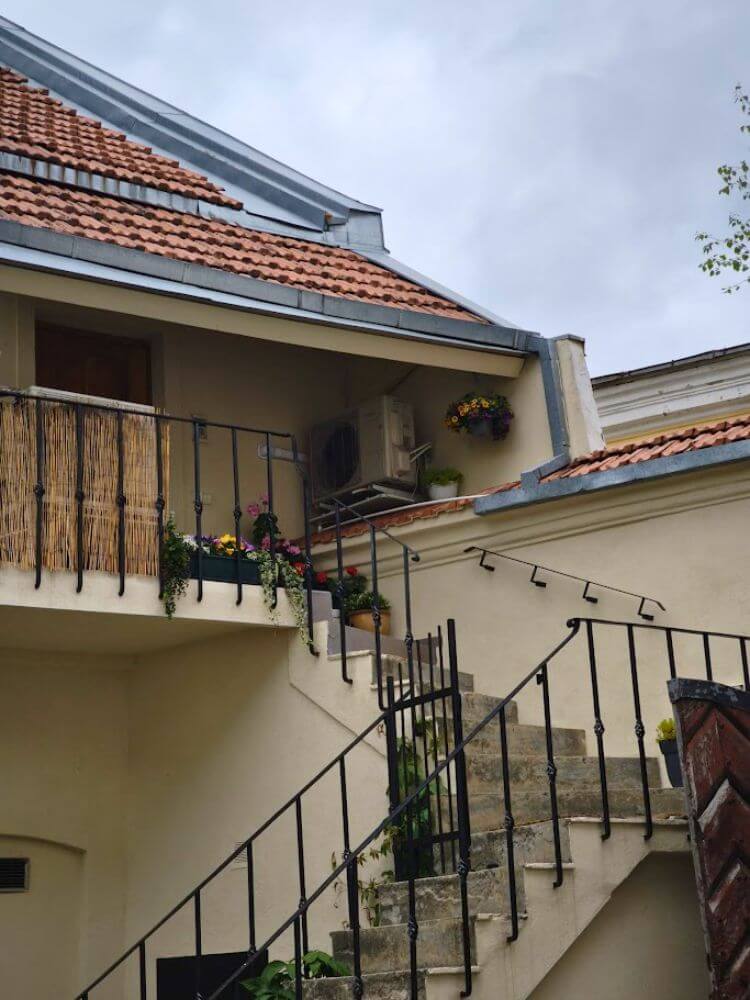 A small Old Town courtyard in Vilnius with outdoor stairs, black railings, flower boxes, and red tile roofs.