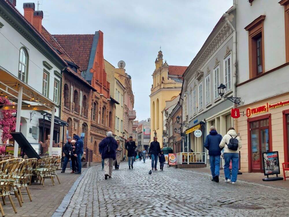 Pilies Street in Vilnius in early April with cobblestones, pedestrians, outdoor café seating, and historic buildings under a gray sky.