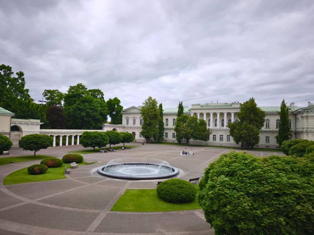 The Presidential Palace courtyard in Vilnius with a round fountain, trimmed greenery, white colonnades, and a cloudy sky.