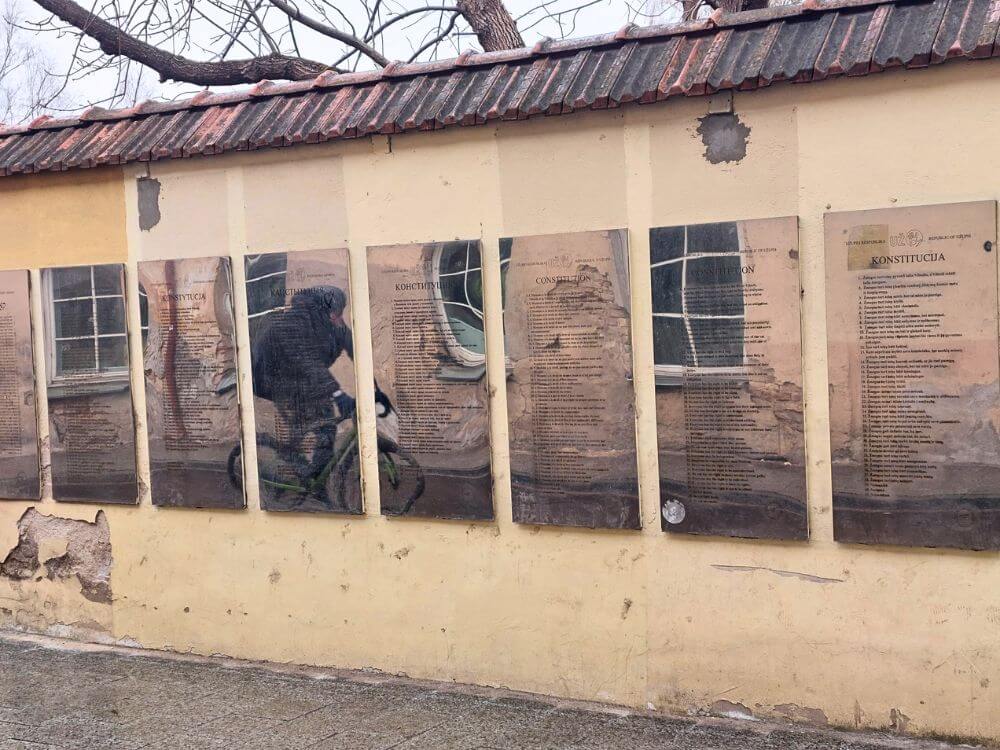 Mirrored plaques of the Užupis Constitution mounted on a yellow wall in Vilnius, with a cyclist reflected in the surface.