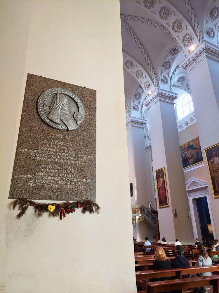 A plaque dedicated to King Mindaugas inside Vilnius Cathedral, with vaulted ceilings, tall columns, paintings, and seated visitors in the background.