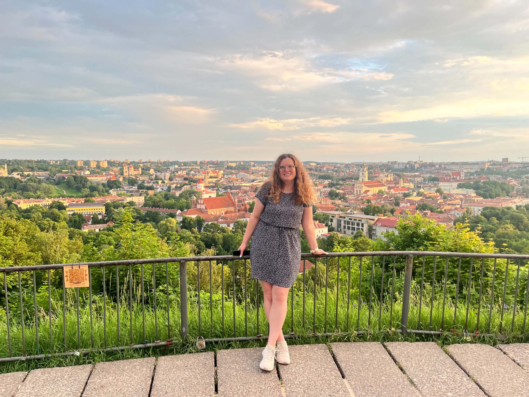 A woman standing at the viewpoint on Three Crosses Hill with the Vilnius panorama, red rooftops, church towers, and greenery behind her